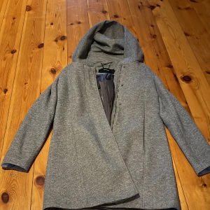 Grå kappa från Vero Moda - Snygg grå kappa från Vero Moda med huva och dragkedja. Perfekt för kyliga dagar med sin stilrena design och praktiska detaljer. Kappan har långa ärmar och en mjuk insida för extra komfort.