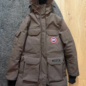 Brun parkajacka från Canada Goose - Säljer en brun parkajacka från Canada Goose med huva och flera praktiska fickor. Jackan har en dragkedja och knappar framtill samt en ikonisk logga på bröstet. Perfekt för kalla dagar. Storlek 2xs