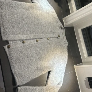 Grå kofta med guldknappar - Snygg grå kofta med dekorativa knappar framtill. Perfekt för en stilren look. Materialet är mjukt och bekvämt, vilket gör den idealisk för både vardag och mer formella tillfällen. Var den är ifrån vet jag inte eftersom att jag fick den av min moster, då den inte hade någon etikett. Den är väl använd och är därför lite nopprig, det är inte något man ser. 