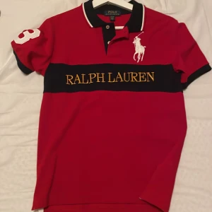 Röd pikétröja från Ralph Lauren - Säljer en snygg röd pikétröja från Ralph Lauren med klassisk krage och knappar. Tröjan har en stor logga på bröstet och nummerdetalj på ärmen. Perfekt för en stilren look. (10-12)