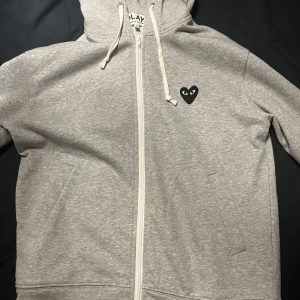 Grå hoodie från Comme des Garçons - Säljer en skön grå hoodie från Comme des Garçons med dragkedja och det ikoniska hjärtmärket på bröstet. Säljer då jag beställde den fast den satt för litet. Passar mer som S än M.