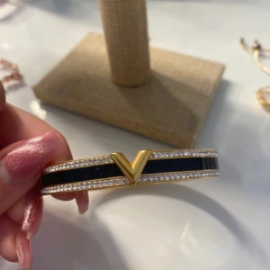 Armband  - Elegant armband i guld med svart emalj och glittrande stenar. Designen har en distinkt V-formad detalj i mitten. Perfekt för att ge en lyxig touch till din stil.