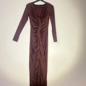 Brun långklänning  - Elegant brun långklänning med v-ringning och volangdetaljer och en slitt framtill. Klänningen har långa ärmar och är tillverkad i ett mjukt, stretchigt material som ger en smickrande passform. Passar XS/S