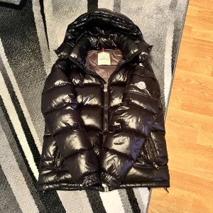 Svart dunjacka från Moncler - Säljer en snygg svart dunjacka från Moncler med glansig finish. Jackan har en huva och dragkedja framtill samt praktiska fickor med dragkedjor. Perfekt för kyliga dagar. Kan sänka vid snabb affär varav att de finns 2 småskador på ärmen