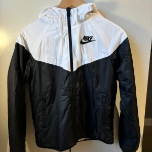Svart och vit vindjacka från Nike - Säljer en stilren svart och vit vindjacka från Nike med dragkedja och huva. Jackan har en klassisk design med Nike-logga på bröstet, perfekt för våren. Pris: 150kr
