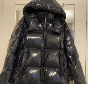 Svart pufferjacka från Tommy Hilfiger - Snygg svart pufferjacka från Tommy Hilfiger med glansig finish och huva. Jackan har en dragkedja framtill och ett diskret märke på ärmen. Perfekt för kyliga dagar.Jackan är endast testad.