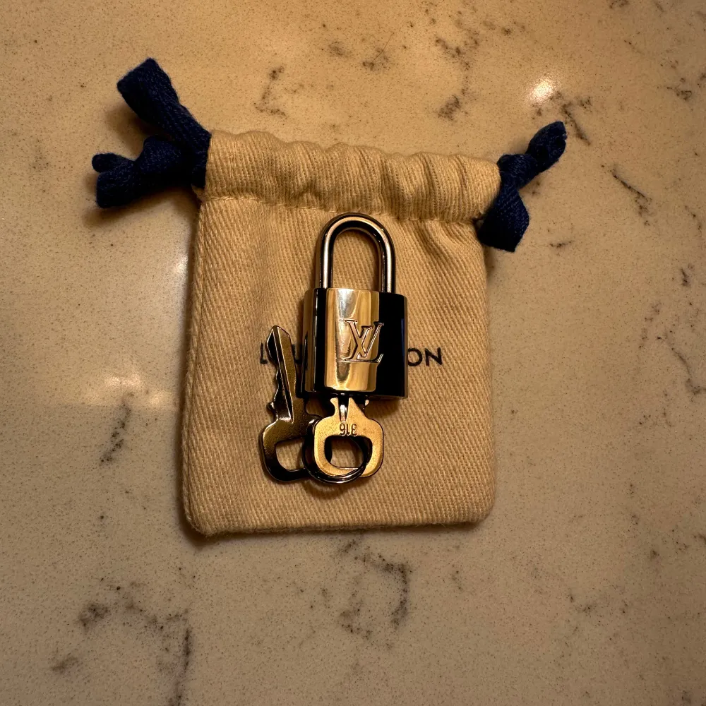 Elegant guld hänglås med tillhörande nycklar från Louis Vuitton. Perfekt för att ge en lyxig touch till din väska eller resväska. Kommer i en snygg liten tygpåse med dragsko.. Asusteet.