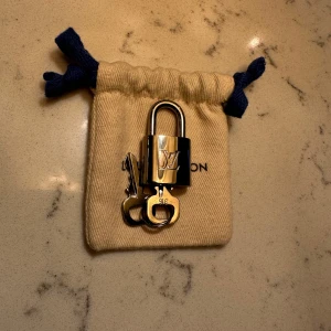 Guld hänglås från Louis Vuitton - Elegant guld hänglås med tillhörande nycklar från Louis Vuitton. Perfekt för att ge en lyxig touch till din väska eller resväska. Kommer i en snygg liten tygpåse med dragsko.