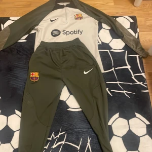 Träningsset från Nike med FC Barcelona-logga - Säljer ett träningsset från Nike med FC Barcelona-logga. Tröjan är grå med långärmade olivgröna ärmar och byxorna är olivgröna. Perfekt för fotbollsträning eller som supporterplagg. Setet har en modern design med klubbens emblem och sponsorlogga.
