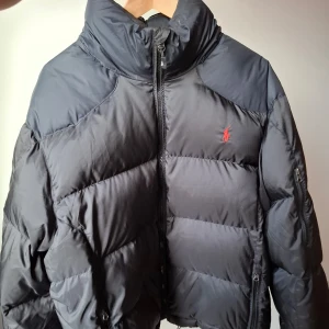 Svart dunjacka från Polo Ralph Lauren - Säljer en svart dunjacka från Polo Ralph Lauren med broderad logga i rött. Jackan har en hög krage och dragkedja framtill. Storlek XXL men sambon som är en XL kunde inte ha den då den satt för tajt och blev även för kort på längden för honom.uppskaytar den som mindre i storlek. I väldigt bra/sparsamt skick! Finns en tunn luva i kragen man kan vika fram vid behov. 