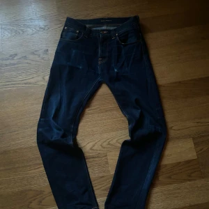 Mörkblå jeans från Nudie Jeans - Säljer ett par mörkblå jeans från Nudie Jeans i ekologisk bomull. De har en klassisk femficksdesign och orange sömmar. Perfekta för en stilren look.