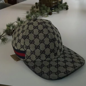 keps Gucci - Snygg keps från Gucci med det ikoniska GG-mönstret i beige och svart. Kepsen har en justerbar rem baktill och en röd och blå randdetalj på sidan. Kepsen luktar lite ciggareter