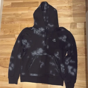 Svart och vit hoodie från Calvin Klein - Säljer en cool svart hoodie från Calvin Klein med vita mönster. Den har en stor huva och en praktisk magficka. Perfekt för en avslappnad stil. Produkten finns ej i sortiment längre! (Passar L)