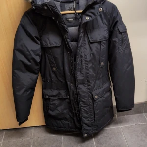 Svart everest parkajacka med huva - Säljer en svart parkajacka från Everest med huva och flera praktiska fickor. Jackan har dragkedja och knappar framtill samt långa ärmar. Perfekt för kyliga dagar. Sparsamt använd 