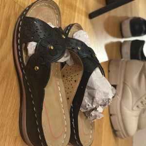 Svarta sandaler med blommönster - Snygga svarta sandaler med dekorativa blommor och guldfärgade detaljer. Perfekta för en avslappnad stil med en touch av elegans.