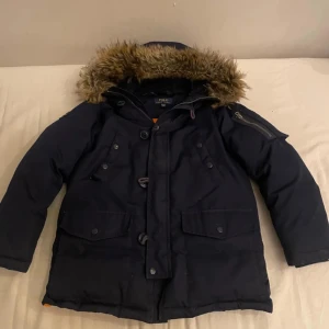 Mörkblå parkajacka med pälsdetaljer - Säljer en snygg mörkblå parkajacka från Ralph lauren. Jackan har en avtagbar huva med pälsdetaljer och knäppning framtill. Perfekt för kalla dagar med sina långa ärmar och praktiska fickor.