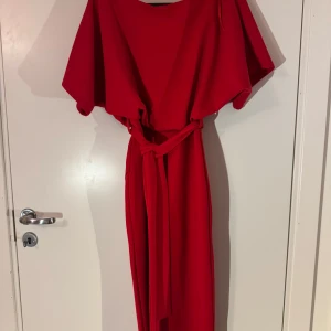Röd klänning från Evita - Snygg röd klänning från Evita med ett bälte i midjan. Klänningen har en elegant omlott-design och korta ärmar. Perfekt för en stilren look.