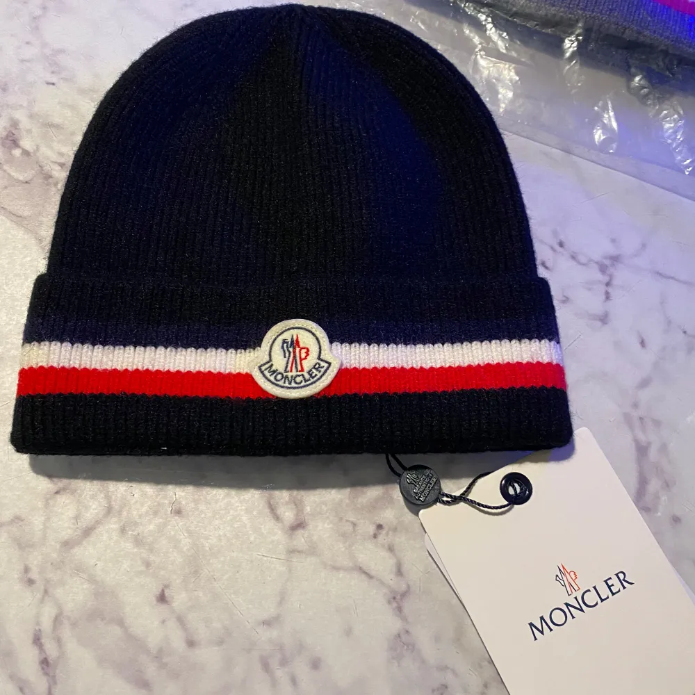 Snygg svart mössa från Moncler med röd och vit rand. Den har Monclers logotyp framtill och är perfekt för kyliga dagar. Mössan är ribbad och ger en stilren look.. Asusteet.