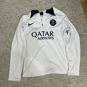 Vit långärmad fotbollströja från Nike - Säljer en vit långärmad fotbollströja från Nike med PSG-logga och Qatar Airways-tryck på framsidan. Tröjan har en dragkedja vid halsen och svarta detaljer vid kragen. Perfekt för fotbollsfans!