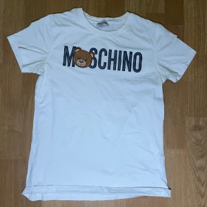 Vit t-shirt från Moschino med björntryck - Säljer en vit t-shirt från Moschino med ett coolt björntryck och texten 'MOSCHINO' på framsidan. Perfekt för en avslappnad stil. T-shirten har en rund hals och korta ärmar.