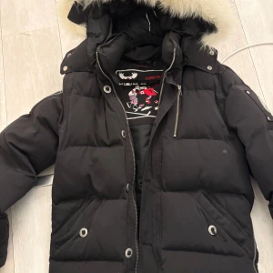 Svart dunjacka från Moose Knuckles - Säljer en svart dunjacka från Moose Knuckles med pälskantad huva. Jackan har dragkedja och knappar framtill samt flera praktiska fickor. Perfekt för kalla vinterdagar.