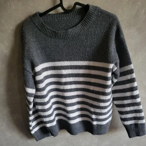 Grå och vit randig stickad tröja - Mysig grå och vit randig stickad tröja med rund hals och långa ärmar. Perfekt för kyliga dagar med sin bekväma passform och klassiska design.