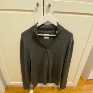 Grå merinoulltröja från These Glory Days - Säljer en stilren grå tröja i merinoull från These Glory Days. Tröjan har en dragkedja framtill och en hög krage, perfekt för kyligare dagar. Den är mjuk och bekväm, idealisk för både vardag och mer uppklädda tillfällen. Nypris 1000 kr