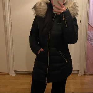 Svart dunjacka från Rockandblue - Snygg svart dunjacka från Rockandblue med pälskantad huva och guldfärgade dragkedjor. Jackan har en quiltad design och är perfekt för kalla dagar. Den har långa ärmar och en figursydd passform.💖