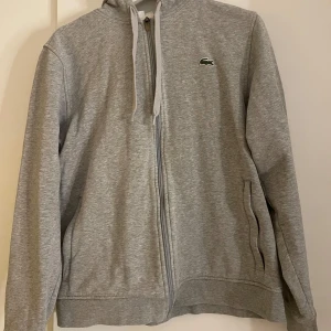 Grå hoodie från Lacoste - Säljer en stilren grå hoodie från Lacoste med dragkedja och justerbar huva. Den har det klassiska krokodilmärket på bröstet och praktiska fickor. 