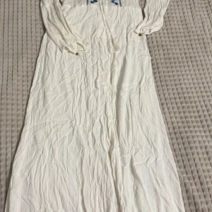 Vit långklänning med broderi - Charmig vit långklänning med blått och svart broderi längs framsidan. Klänningen har långa ärmar och ett knytband i midjan för en justerbar passform. Perfekt för en bohemisk look. Om ni vill se den på, skriv gärna!