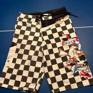Ed Hardy shorts - Coola svartvita rutiga shorts med ett stort dödskallemotiv och färgglada detaljer. Shortsen har en praktisk ficka med knapp och snörning i midjan för justerbar passform. Perfekta för en avslappnad stil med en edgy touch.