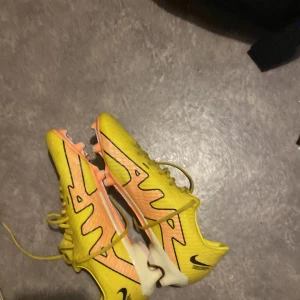 Gula fotbollsskor från Nike air  - Säljer ett par gula Nike fotbollsskor med svarta detaljer och snörning. Skorna har en orange sula med dobbar, perfekt för gräsplaner. De är designade för att ge bra grepp och komfort under spelet.