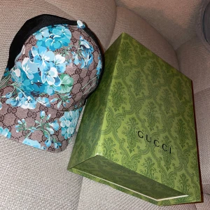 Blommig keps från Gucci - Snygg keps från Gucci med ett blommigt mönster i blått på en brun bas. Kepsen har justerbar passform och märkeslogga på insidan. Perfekt för att ge en färgklick till din outfit!