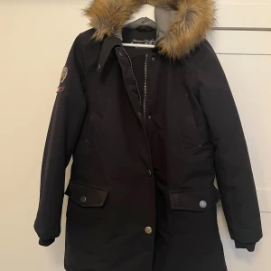 Svart parkajacka med päls från Berkeley - Säljer en svart parkajacka från Berkeley med en mysig pälsfodrad huva. Jackan har knappar och dragkedja framtill samt praktiska fickor med knappar. Perfekt för kalla dagar!