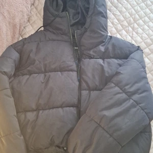 Svart pufferjacka från Monki - Säljer en svart croppad pufferjacka från Monki i storlek M. Jackan är perfekt för kalla dagar med sin vadderade design och har en praktisk dragkedja framtill. Den har en huva för extra skydd mot väder och vind. Jackan är i bra skick.  