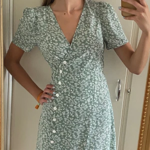 Blommig mintgrön omlottklänning - Mintgrön omlottklänning med blommönster i vitt, v-ringning och puffärmar. Ungefär midi-längd (slutar en bit över knäet) med tunt och bekvämt material. Storlek är S. Köpt second hand. Perfekt sommarklänning!