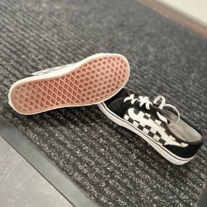 Svartvita rutiga sneakers - Säljer ett par coola svartvita vans sneakers med klassiskt rutigt mönster. Skorna har en vit sula med en brun undersida och snörning framtill. Perfekta för en avslappnad stil!