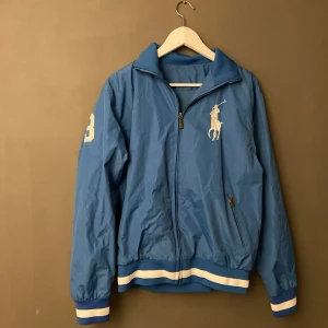Blå vindjacka från Ralph Lauren - Säljer en snygg blå vindjacka från Ralph Lauren. Jackan har en dragkedja framtill och är dekorerad med det klassiska Ralph Lauren-logotypen på bröstet. Den har vita och blå ränder vid ärmslut och nederkant, samt en siffra på ärmen. Perfekt för vår och höst!