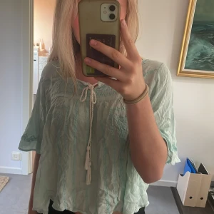 Ljusgrön blus med knytband - Säljer en söt ljusgrön blus med knytband framtill. Den har korta ärmar och en lös passform, perfekt för varma dagar. Blusen är i ett lätt och luftigt material, vilket gör den superbekväm att bära.