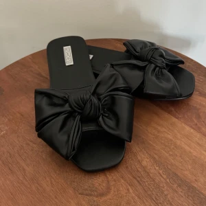 Svarta sandaler med rosett - Säljer ett par snygga svarta sandaler med en stor rosett på ovansidan. Perfekta för sommaren och passar till både vardag och fest. De är i nyskick och väldigt bekväma att ha på sig.