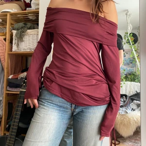 Vinröd offshoulder topp - Säljer en snygg vinröd offshoulder topp i ett mjukt och stretchigt material. Toppen har långa ärmar och en draperad design som ger en cool och avslappnad look.