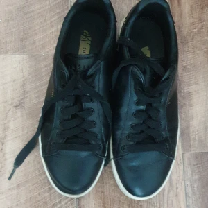 Svarta sneakers från SikSilk - Säljer ett par svarta sneakers från SikSilk. De har en klassisk design med snörning och en vit sula. Skorna är i skinn och har en diskret logga på sidan. Perfekta för vardagsbruk!