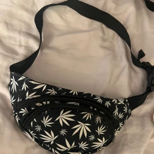 Svart midjeväska med vita bladmotiv - Säljer en cool svart midjeväska med vita bladmotiv. Väskan har två dragkedjefack och en justerbar rem för perfekt passform. Perfekt för festivaler eller vardagsbruk!