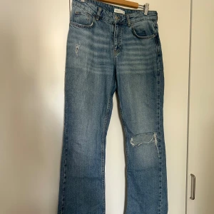 Blå jeans med slitningar - Säljer ett par snygga blå jeans från Gina Tricot i storlek 38. De har en cool slitning på ena benet och en klassisk femficksdesign. Hårt material.