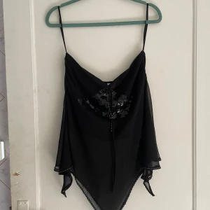 Svart ärmlös topp med spetsdetaljer - Säljer en elegant svart ärmlös topp med tunna axelband och spetsdetaljer. Toppen har en vacker snörning framtill och är perfekt för festliga tillfällen. Materialet är lätt och luftigt, vilket gör den bekväm att bära.