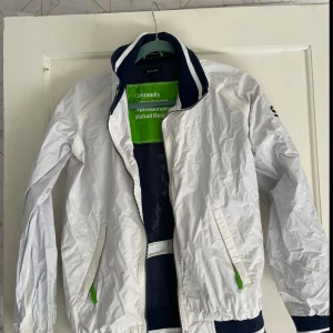 Vit vindjacka från GANT - Säljer en snygg vit vindjacka från GANT i storlek M. Jackan har en sportig look med gröna detaljer och en dragkedja framtill. Perfekt för vår och höst med sin lätta och bekväma passform. Den har långa ärmar och två praktiska fickor med dragkedja.