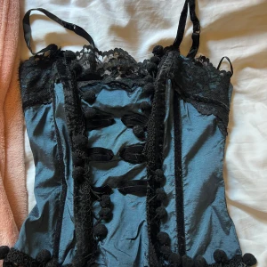 Svart och blå korsett med spetsdetaljer - Säljer en snygg korsett i svart och blått med fina spetsdetaljer. Den har justerbara axelband och en vacker spetskant upptill. Framsidan är dekorerad med knappar och spetsband som ger en elegant look. Perfekt för fest eller speciella tillfällen!