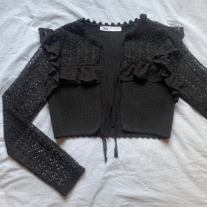 Svart stickad kofta från Zara - Säljer en superfin svart stickad kofta från Zara. Den har långa ärmar med spetsdetaljer och volanger på framsidan. Koftan är croppad och har en knytning framtill. Nypris 360, använd en gång 🖤
