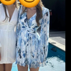 Blommig klänning med långa ärmar - Snygg kortklänning med blommigt mönster i blå och vita toner. Klänningen har en omlott-design med v-ringning och långa. Storlek 34 från NA-KD. Använd endast 1 gång. 