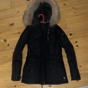 Svart parkajacka med pälsdetalj - Snygg svart parkajacka med avtagbar pälsdetalj på huvan.Den är från Hollie’s  och har använt den ett par gånger, fortfarande lika fin. Jackan har dragkedja och knappar framtill samt flera praktiska fickor. Perfekt för kyliga dagar. ( äckta päls )